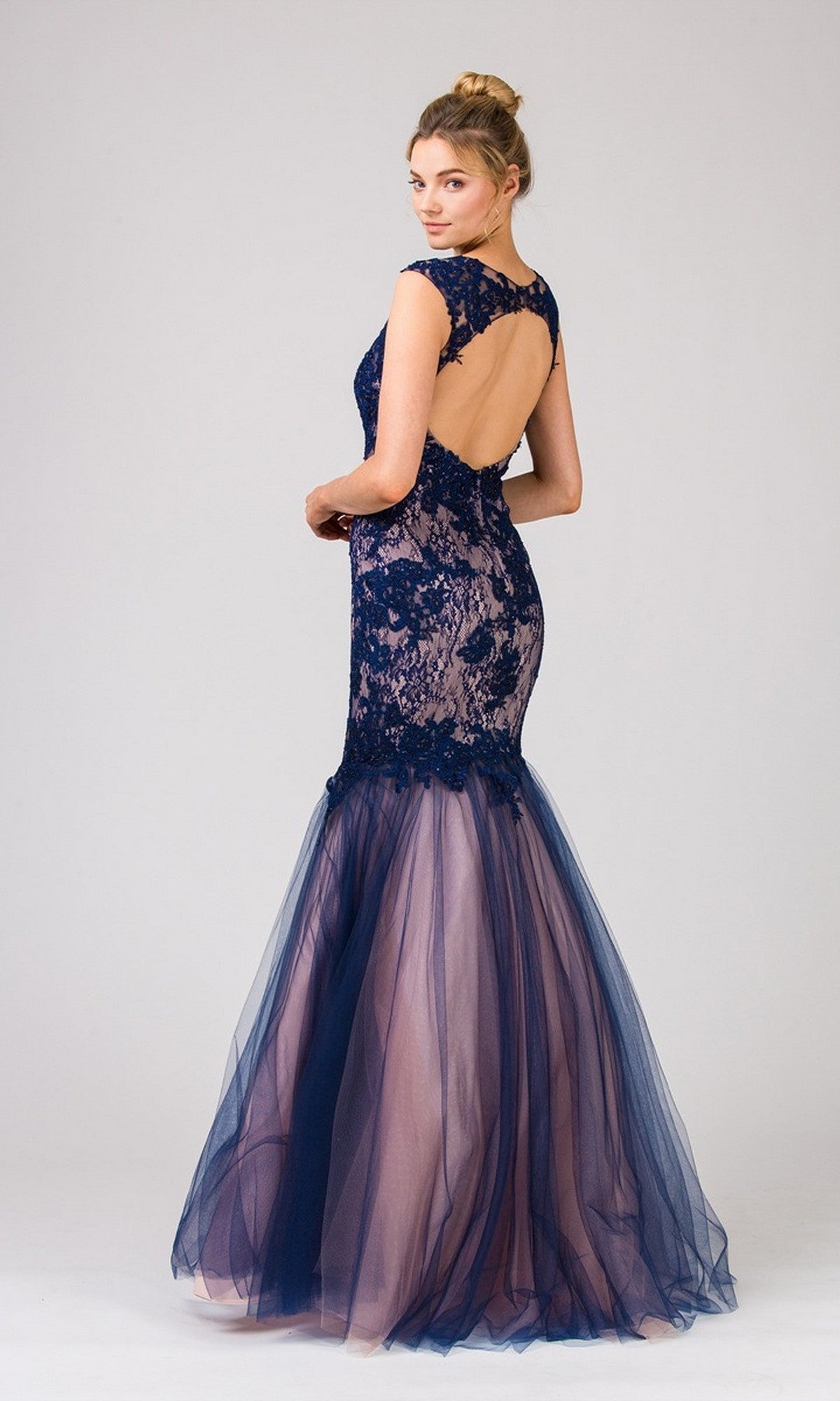 A woman wearing the Long Prom Dress: Eureka 8510โa navy blue lace mermaid gown with a sleeveless bodice, sheer tulle skirt, and open backโfaces away from the camera against a plain background.